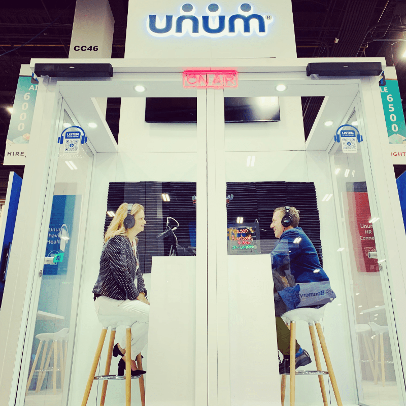 A woman and man are seated across from each other on stools in a glass sound booth with microphones in front of them. The Unum logos appears above the booth.