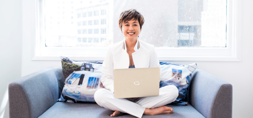 Jenn Lim smiles sitting cross-legged on a couch holding a laptop in front of her.