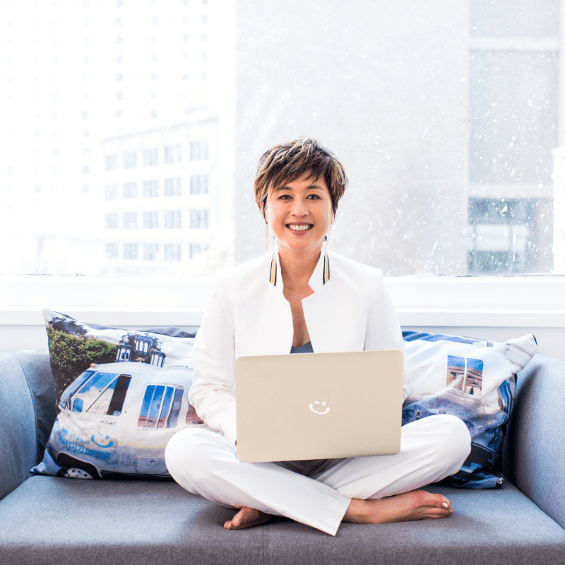 Jenn Lim smiles sitting cross-legged on a couch holding a laptop in front of her.