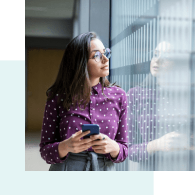 A woman looks out the window of hallway while holding a mobile device. The woman's reflection in the window is visible.