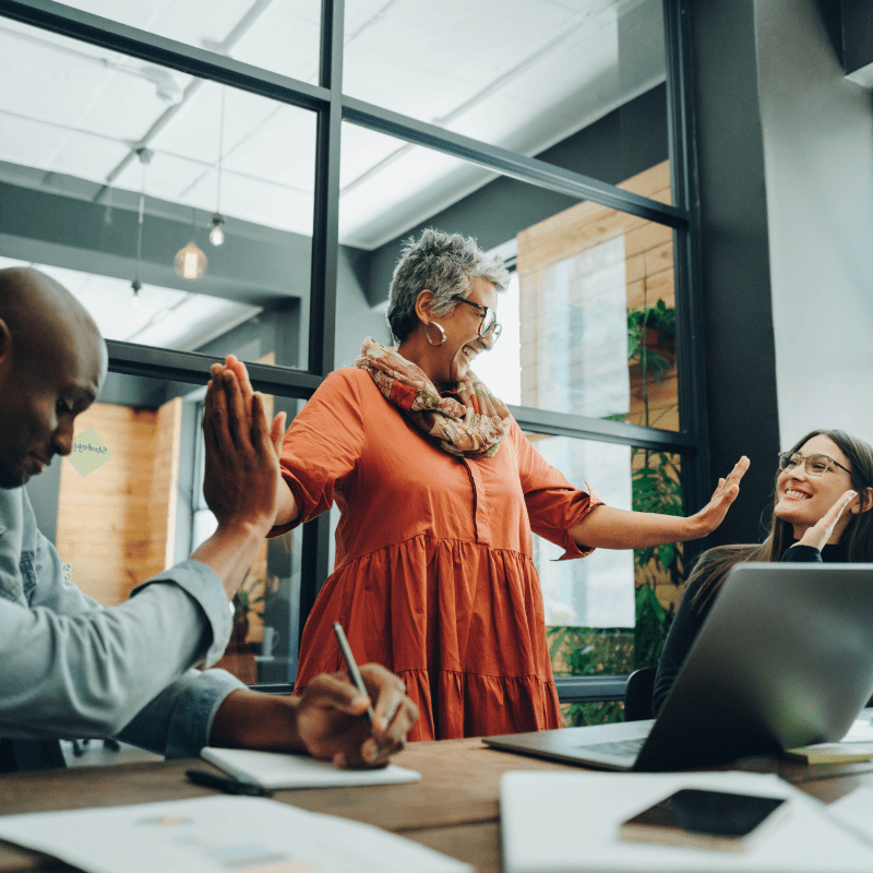 An older woman smiles while standing at a meeting room table with her arms extended to a high-five gesture toward a younger black man and white woman seated on each side of her.