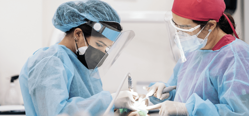 Two women dressed in dental worker gowns and wearing face masks and face shields while working on a patient. One of the women is holding a dental instrument as the other looks on.