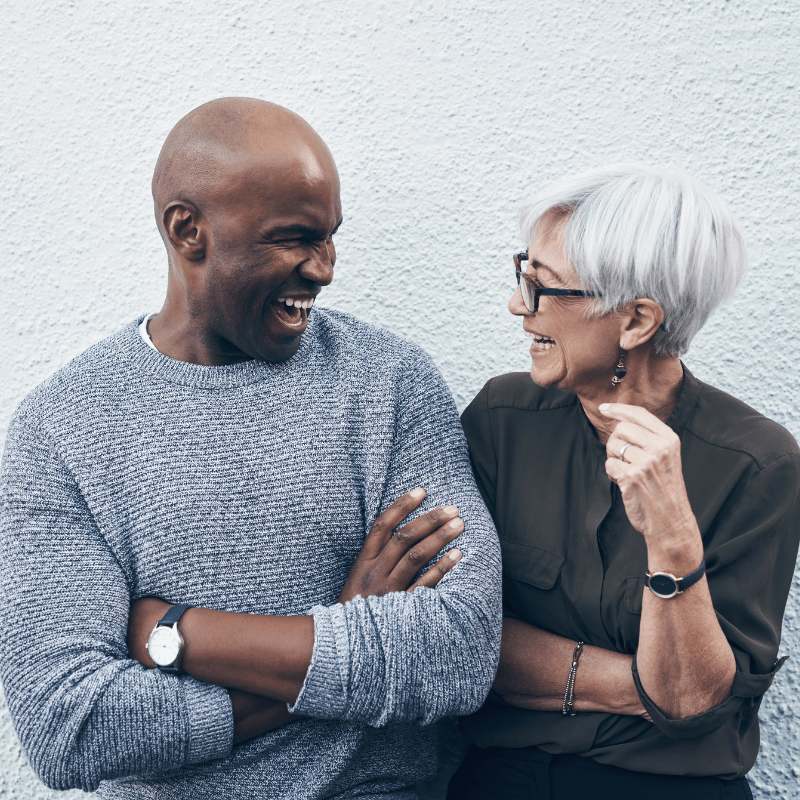 A black man and an older white woman laugh together while standing next to a wall.
